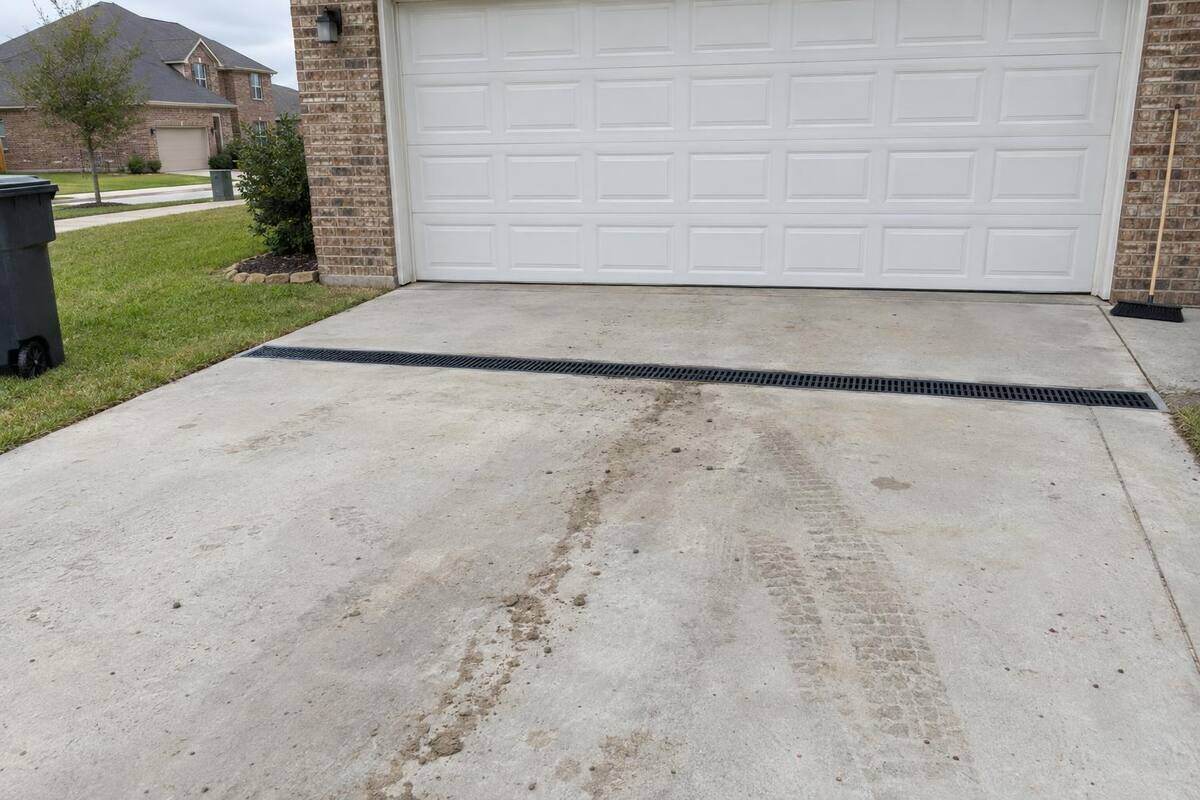A slotted channel drain cut across a concrete driveway just in front of a white garage door